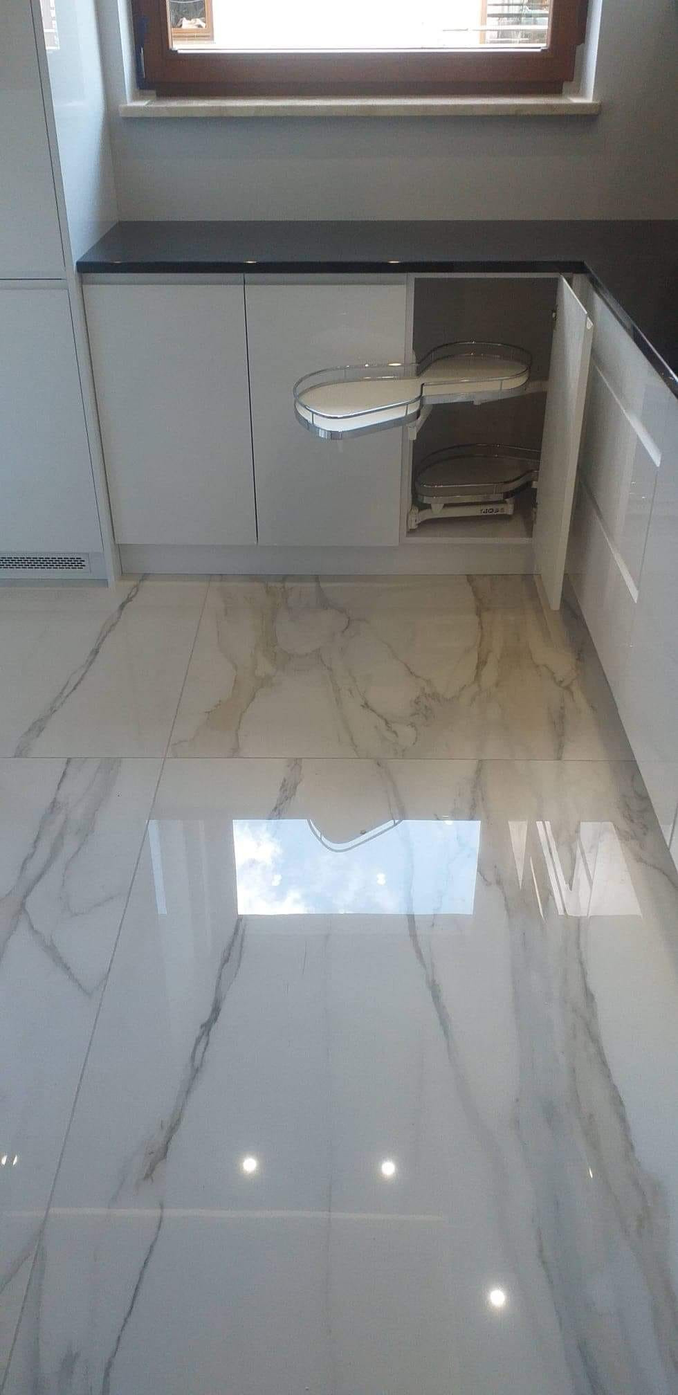 Nowoczesna kuchnia z białymi szafkami i obrotowymi półkami w narożnej szafce, widoczne marmurowe płytki podłogowe z odbiciem okna.