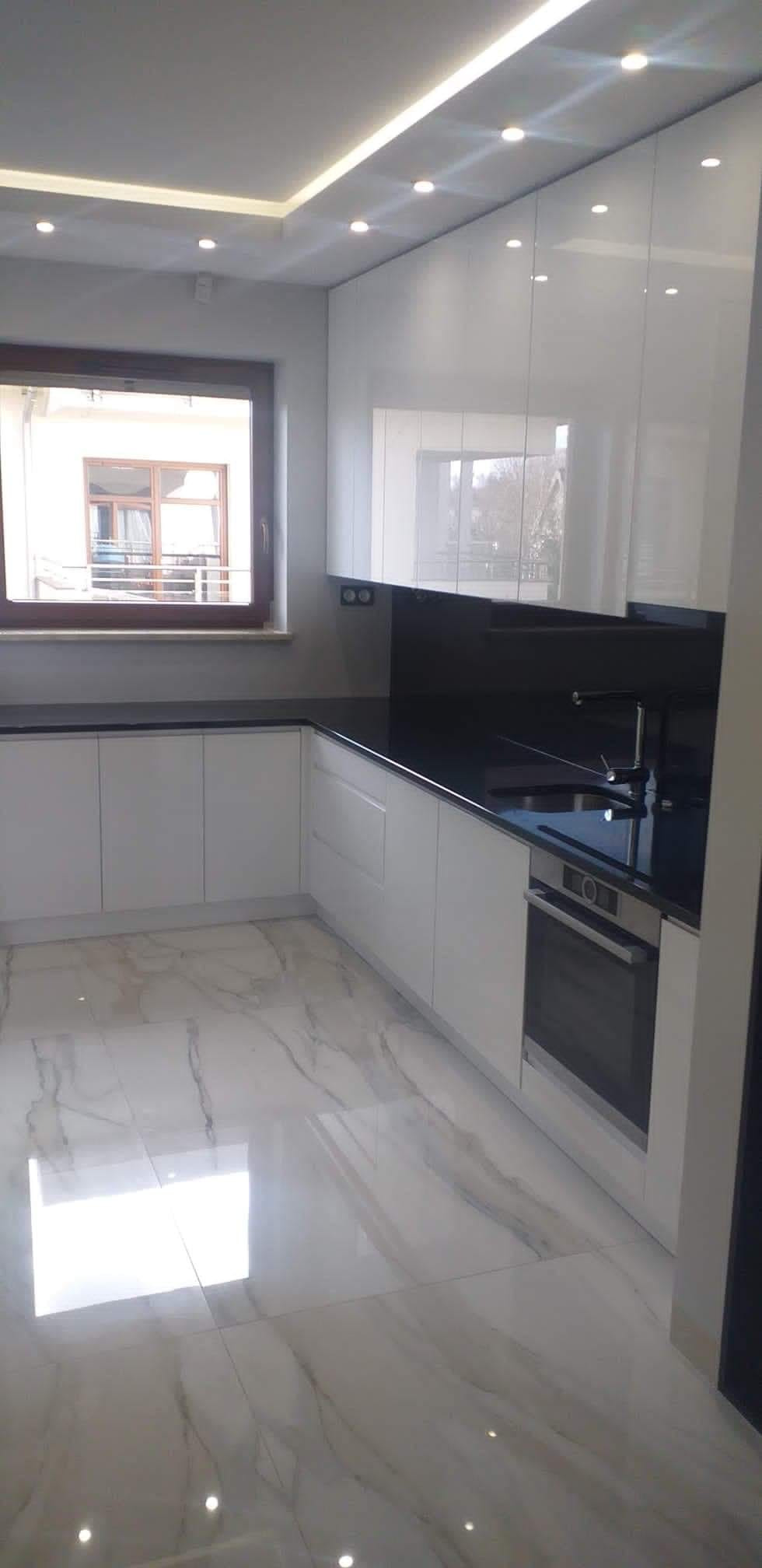 Nowoczesna kuchnia z białymi, lakierowanymi szafkami, czarnym blatem i marmurową podłogą odbijającą światło punktowych lamp sufitowych.