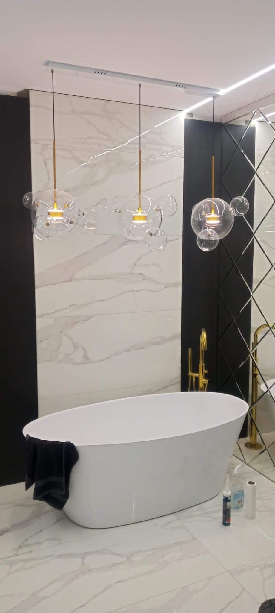 Nowoczesna łazienka z owalną wanną, marmurowymi płytkami na ścianach i podłodze, oraz trzema designerskimi lampami wiszącymi z przezroczystymi kloszami w kształcie bąbelków, czarny ręcznik...