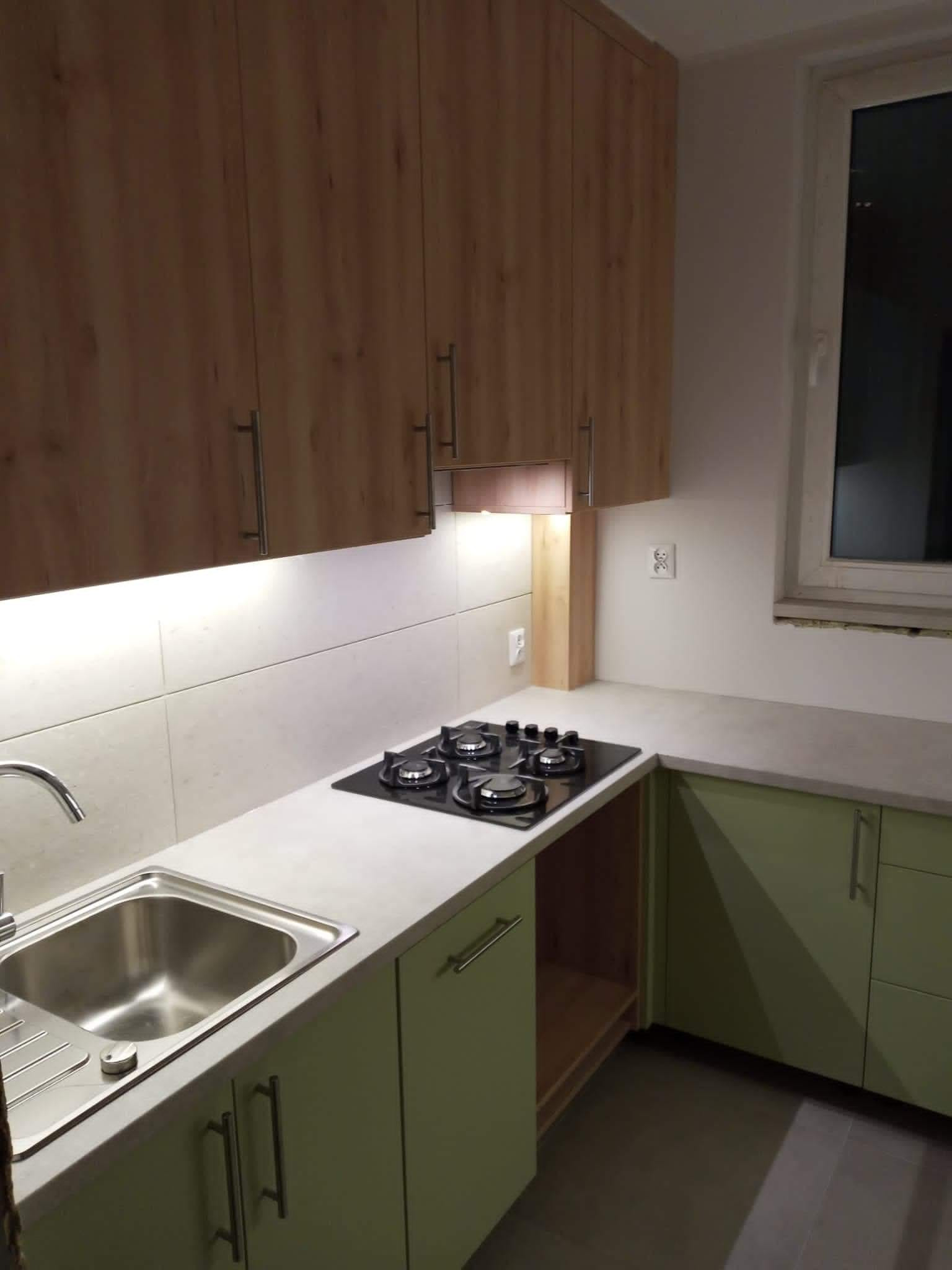 Kuchenna zabudowa: górne szafki z drewnopodobnym fornirem, dolne szafki w odcieniach zieleni, jasny blat z wbudowaną płytą gazową i zlewozmywakiem, oświetlenie podszafkowe LED.