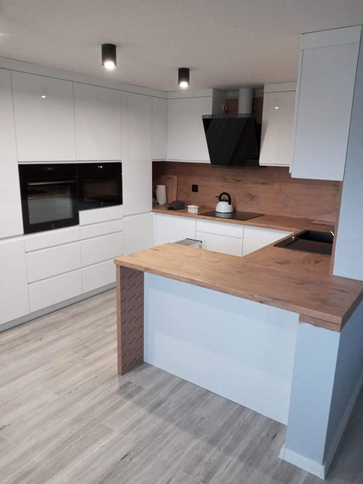 Nowoczesna kuchnia z białymi, lakierowanymi szafkami, drewnianym blatem i okładziną ścienną, czarnym okapem i szarymi płytkami podłogowymi imitującymi drewno.