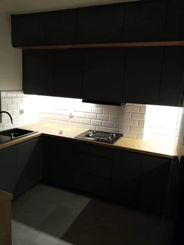 Nowoczesna, ciemna kuchnia z drewnianym blatem, białymi kafelkami metra i oświetleniem LED pod szafkami.