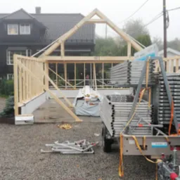 Wznoszenie drewnianej konstrukcji szkieletowej budynku, widoczne rusztowania Layher na przyczepce i materiały budowlane, w tle dom z ciemną elewacją i szarym dachem w mglisty dzień.