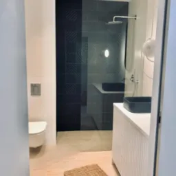 Nowoczesna łazienka z kabiną prysznicową wyłożoną ciemnymi płytkami z geometrycznym wzorem, jasnymi drewnianymi podłogami, białą umywalką z ciemną nablatową misą i dywanikiem z naturalnego włókna