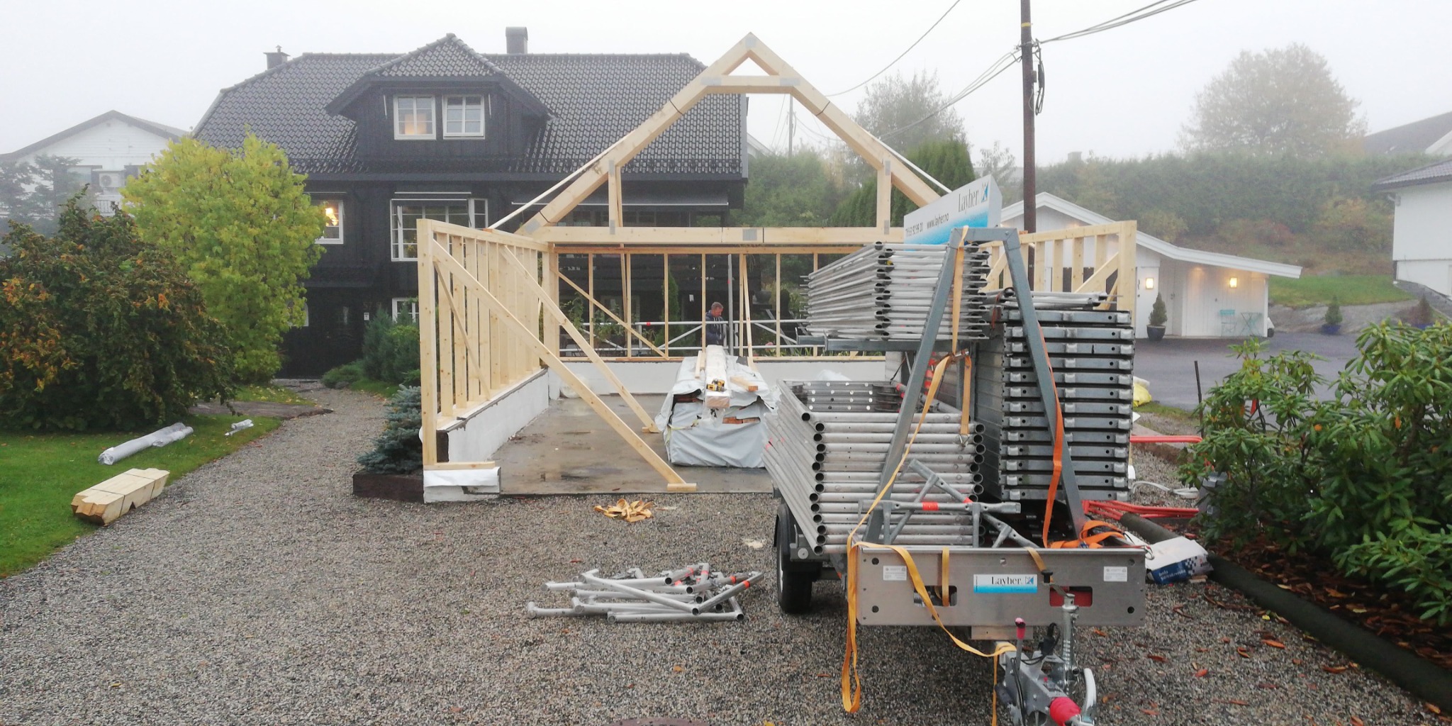 Wznoszenie drewnianej konstrukcji szkieletowej budynku, widoczne rusztowania Layher na przyczepce i materiały budowlane, w tle dom z ciemną elewacją i szarym dachem w mglisty dzień.