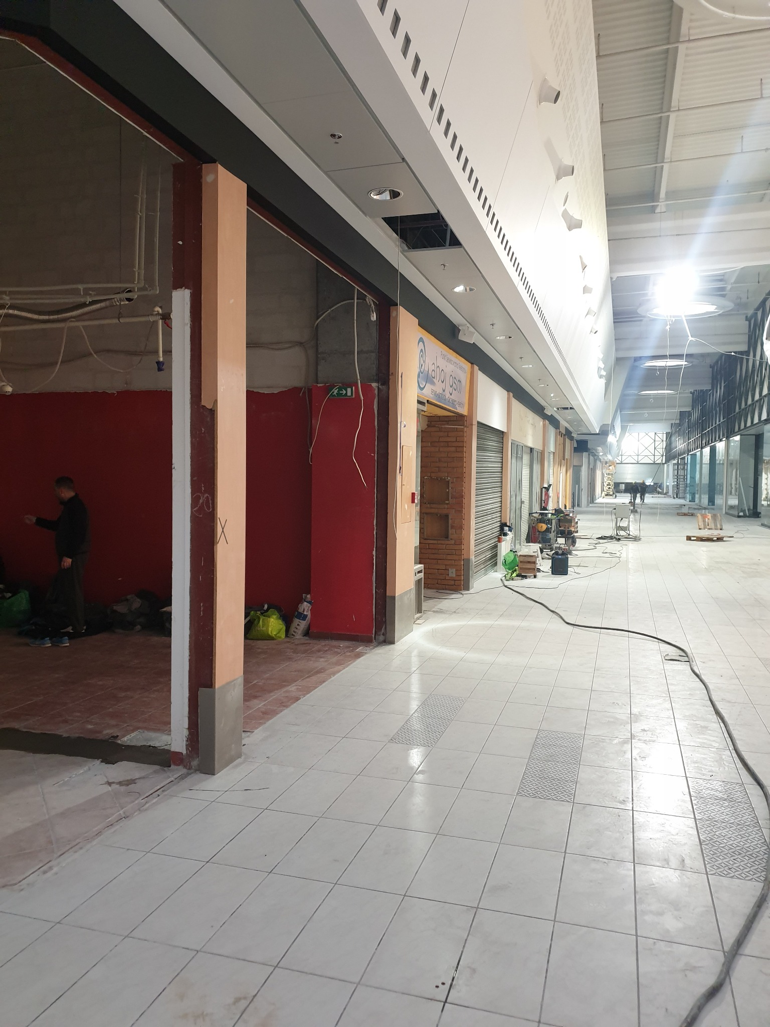 Wnętrze pasażu handlowego w trakcie remontu: widoczne otwarte lokale w surowym stanie, podłoga z płytek w jasnych odcieniach, częściowo zakryta folią ochronną,  pracownik w oddali i porozrzucane...