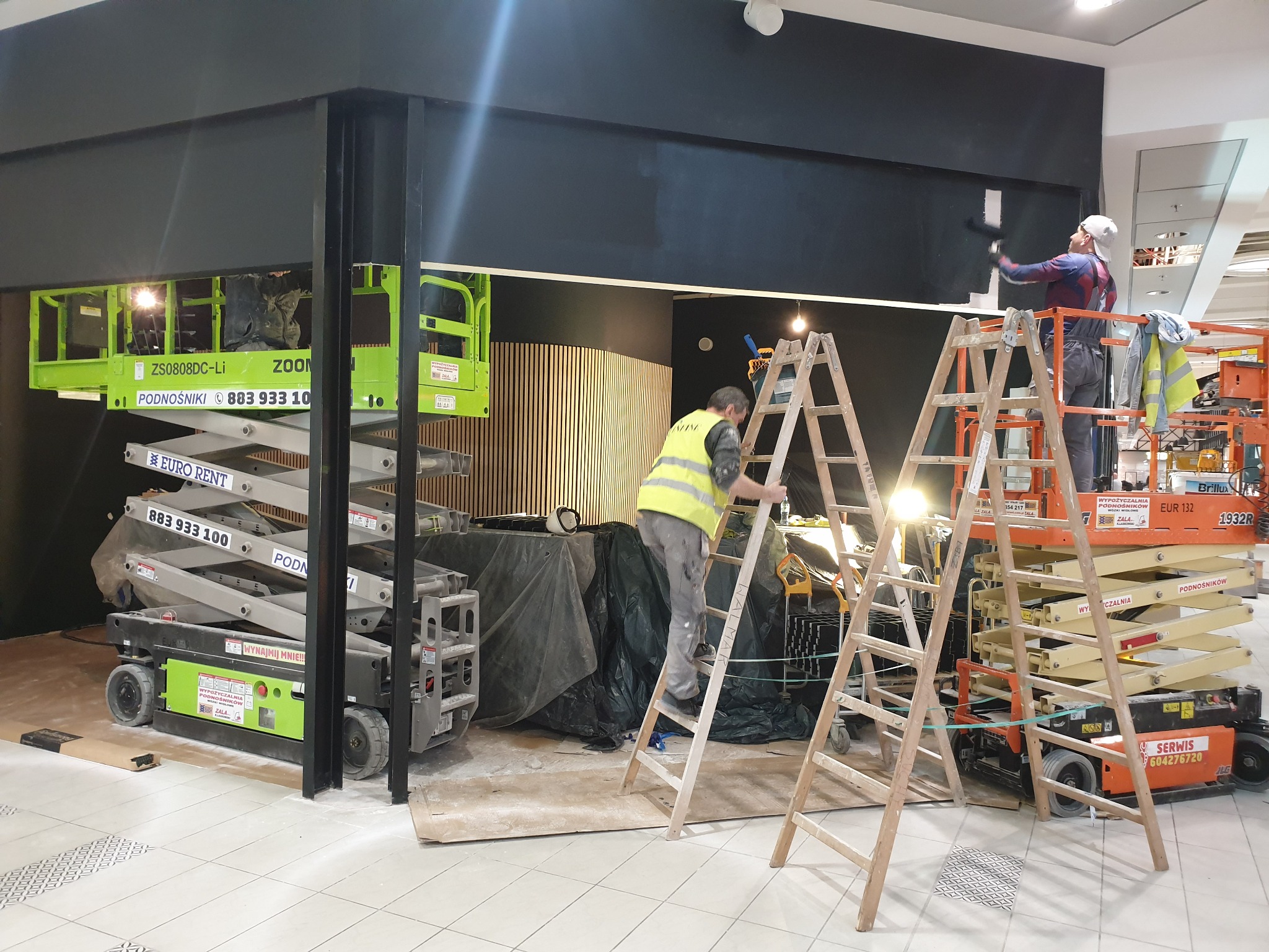 Dwóch pracowników maluje czarną elewację sklepu w galerii handlowej, używając podnośników i drabiny, widoczne narzędzia i osłonięte elementy wnętrza.