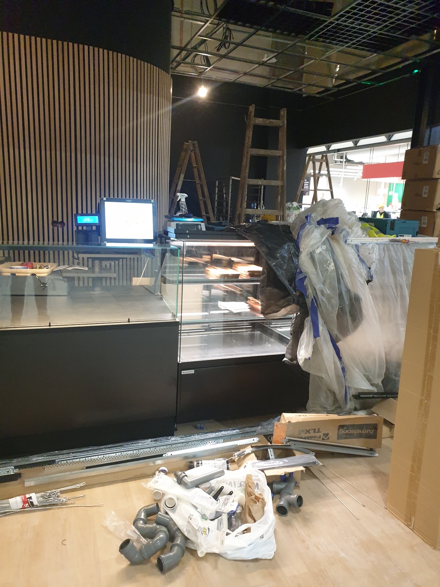 Wnętrze lokalu w trakcie wykańczania: widoczna lada chłodnicza z komputerem, drewniana ściana z pionowych listew, drabiny i materiały budowlane, w tym rury PCV i kartony.