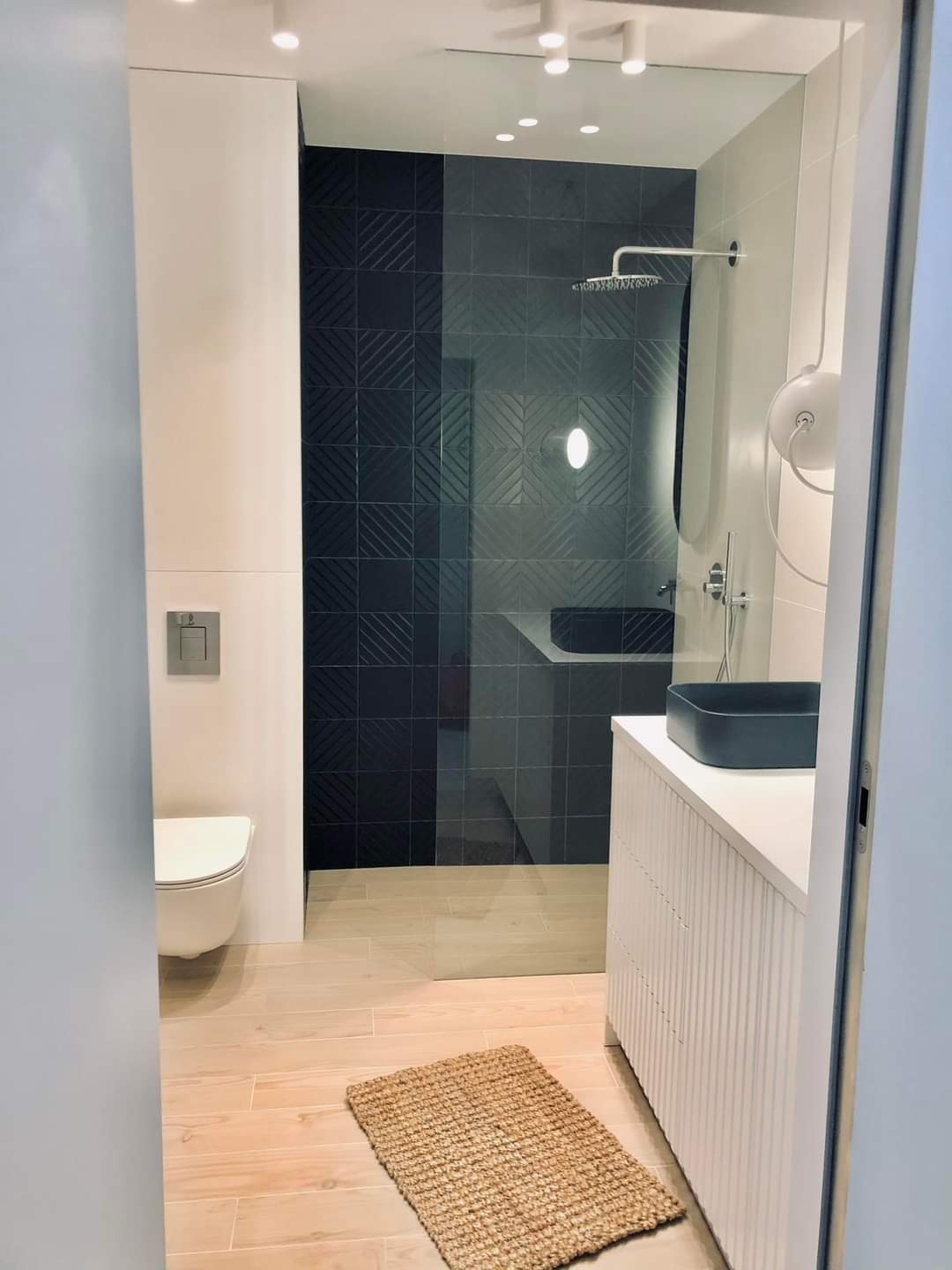 Nowoczesna łazienka z kabiną prysznicową wyłożoną ciemnymi płytkami z geometrycznym wzorem, jasnymi drewnianymi podłogami, białą umywalką z ciemną nablatową misą i dywanikiem z naturalnego włókna