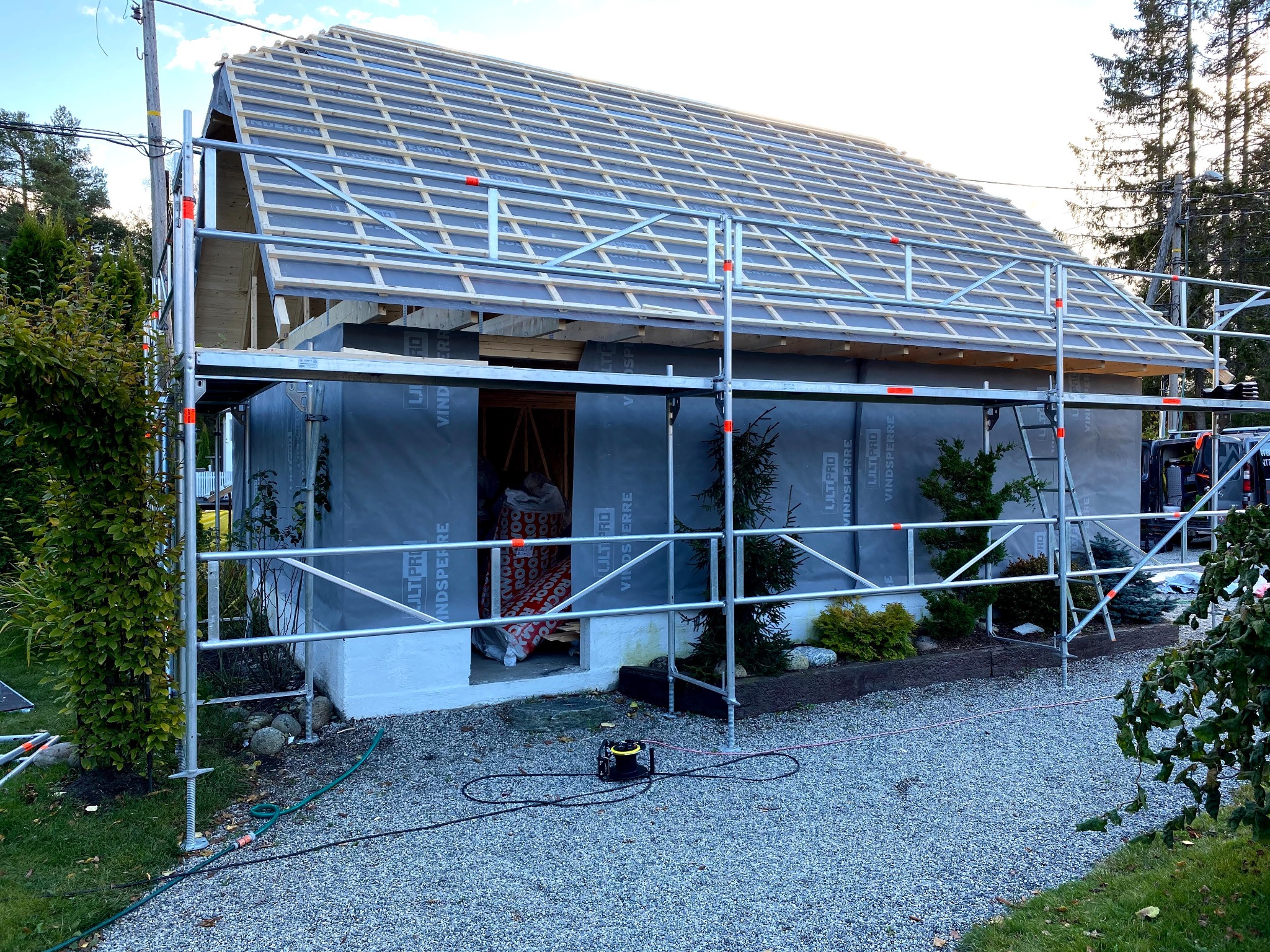 Budynek w trakcie ocieplania, widoczne rusztowania, membrana wiatroizolacyjna na ścianach, drewniana konstrukcja dachu w trakcie budowy, narzędzia na ziemi.