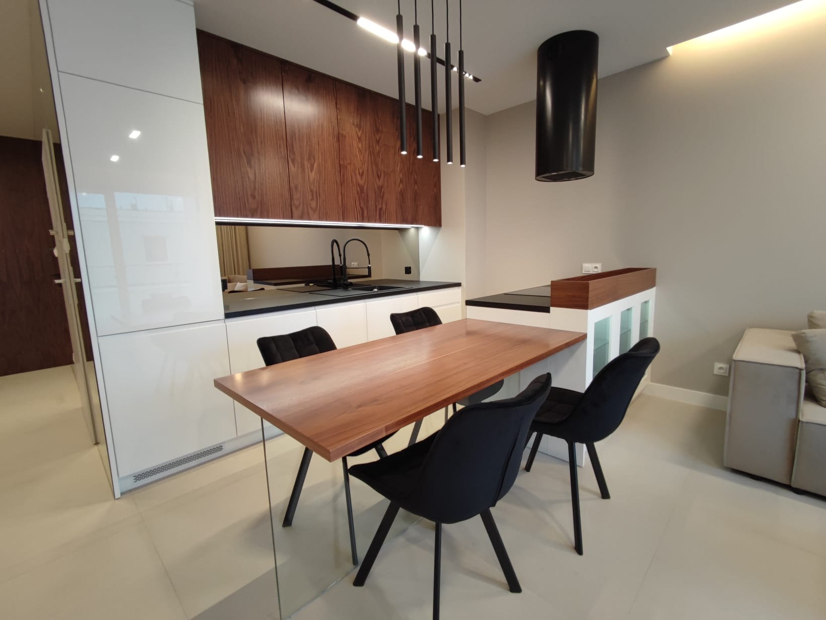 Nowoczesna kuchnia z drewnianymi szafkami, czarnym okapem i stołem z drewnianym blatem oraz czarnymi krzesłami na tle jasnej podłogi.