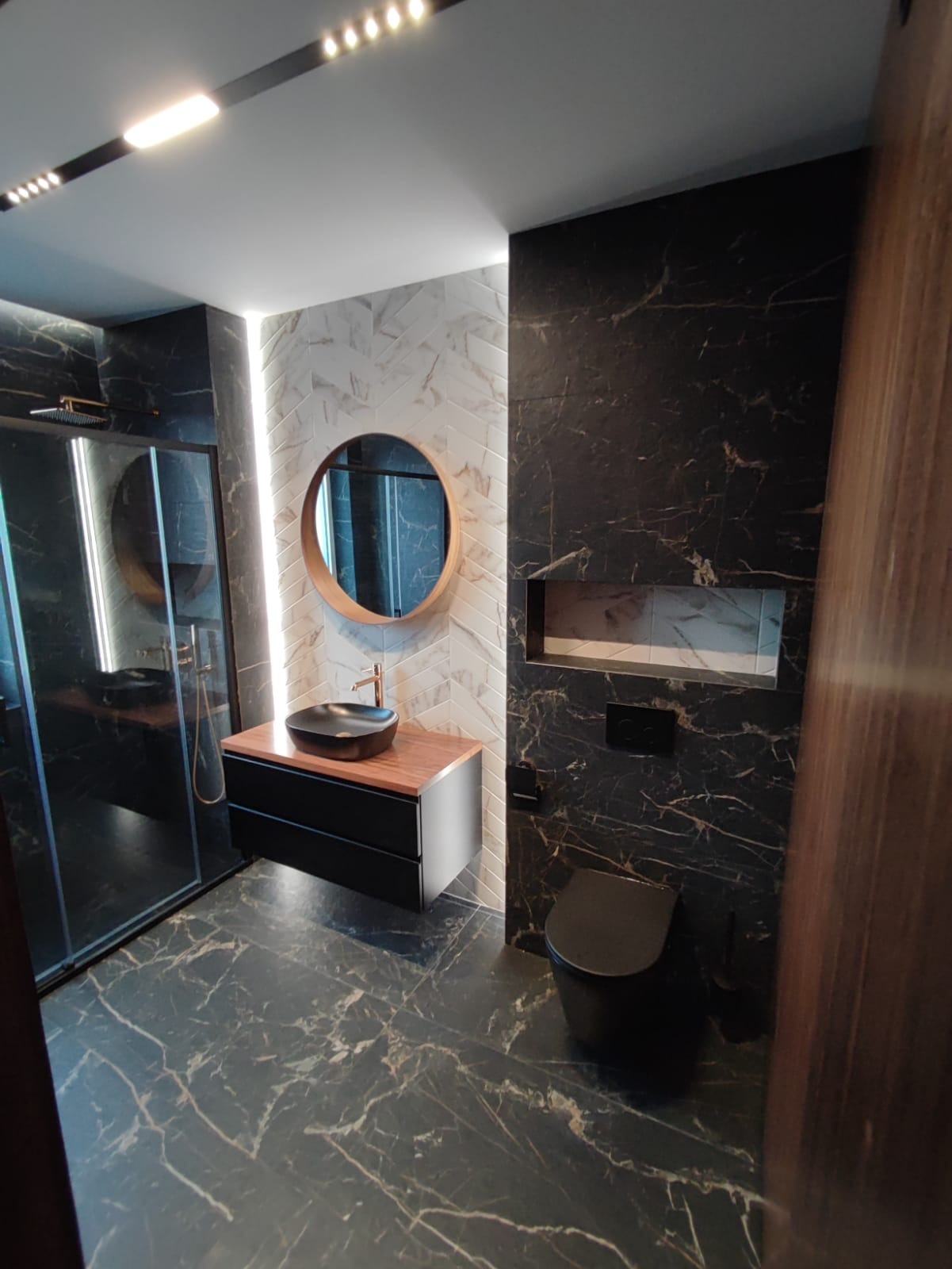 Nowoczesna łazienka z czarnymi marmurowymi płytkami, okrągłym lustrem z drewnianą ramą, czarną umywalką nablatową z miedzianą baterią i wnęką w ścianie.