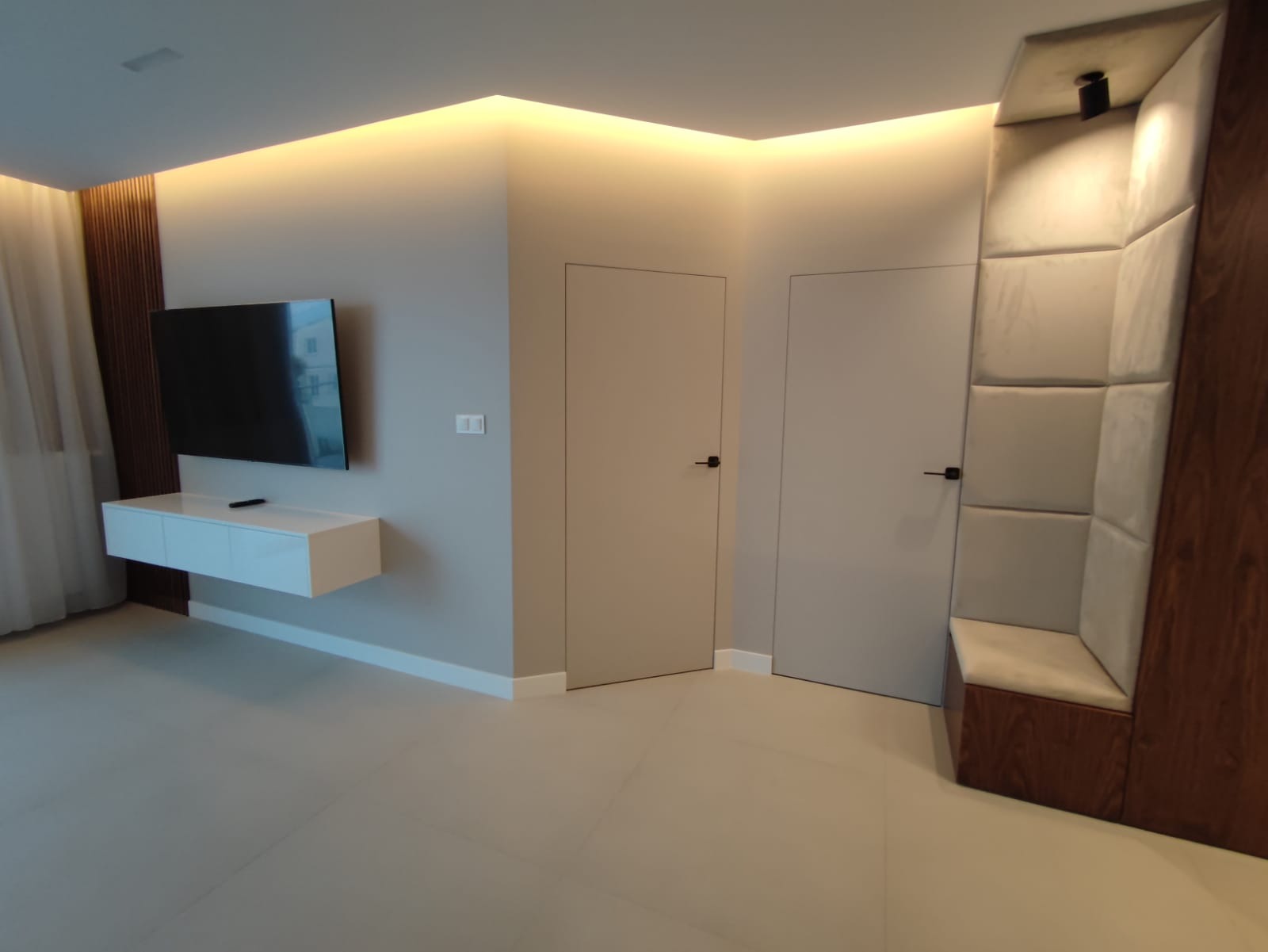 Minimalistyczne wnętrze: telewizor na ścianie, biała szafka RTV, dwie ukryte drzwi w kolorze ścian, dekoracyjny panel ścienny z siedziskiem, podświetlenie LED sufitowe.