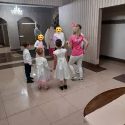 Grupa dzieci w eleganckich strojach, z tyłu animator w różowej koszulce z napisem, w przestronnym, oświetlonym holu z żyrandolami, prawdopodobnie podczas przyjęcia.