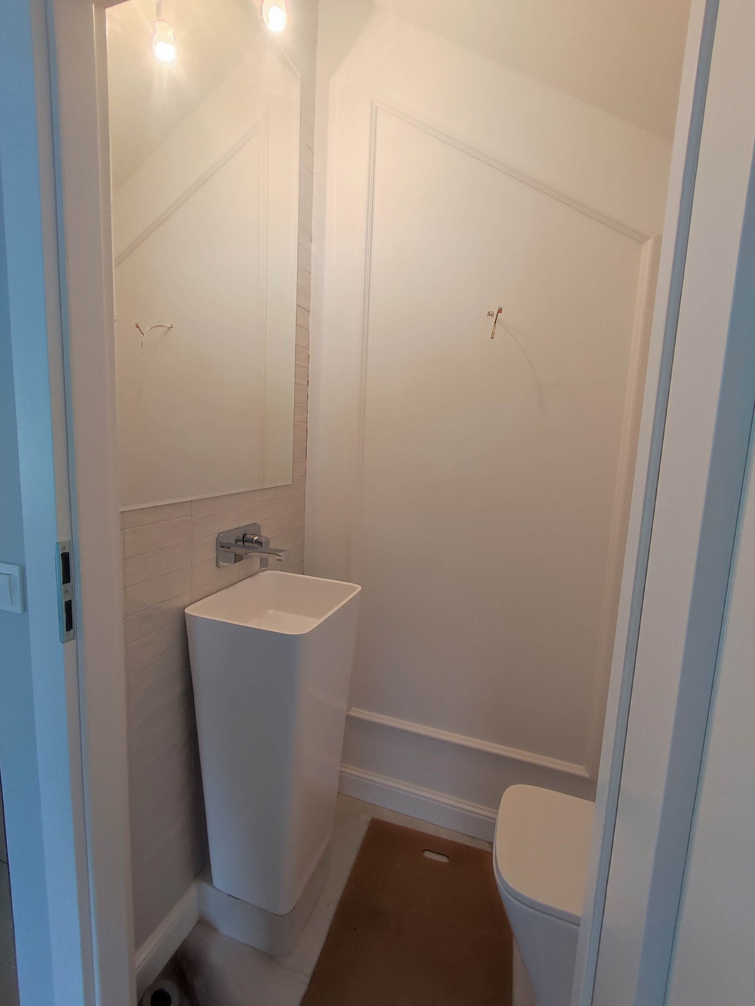 Biała, minimalistyczna umywalka kolumnowa z chromowaną baterią, zamontowana w małej łazience z białymi ścianami wykończonymi listwami i płytkami imitującymi cegłę, widok zza otwartych drzwi.