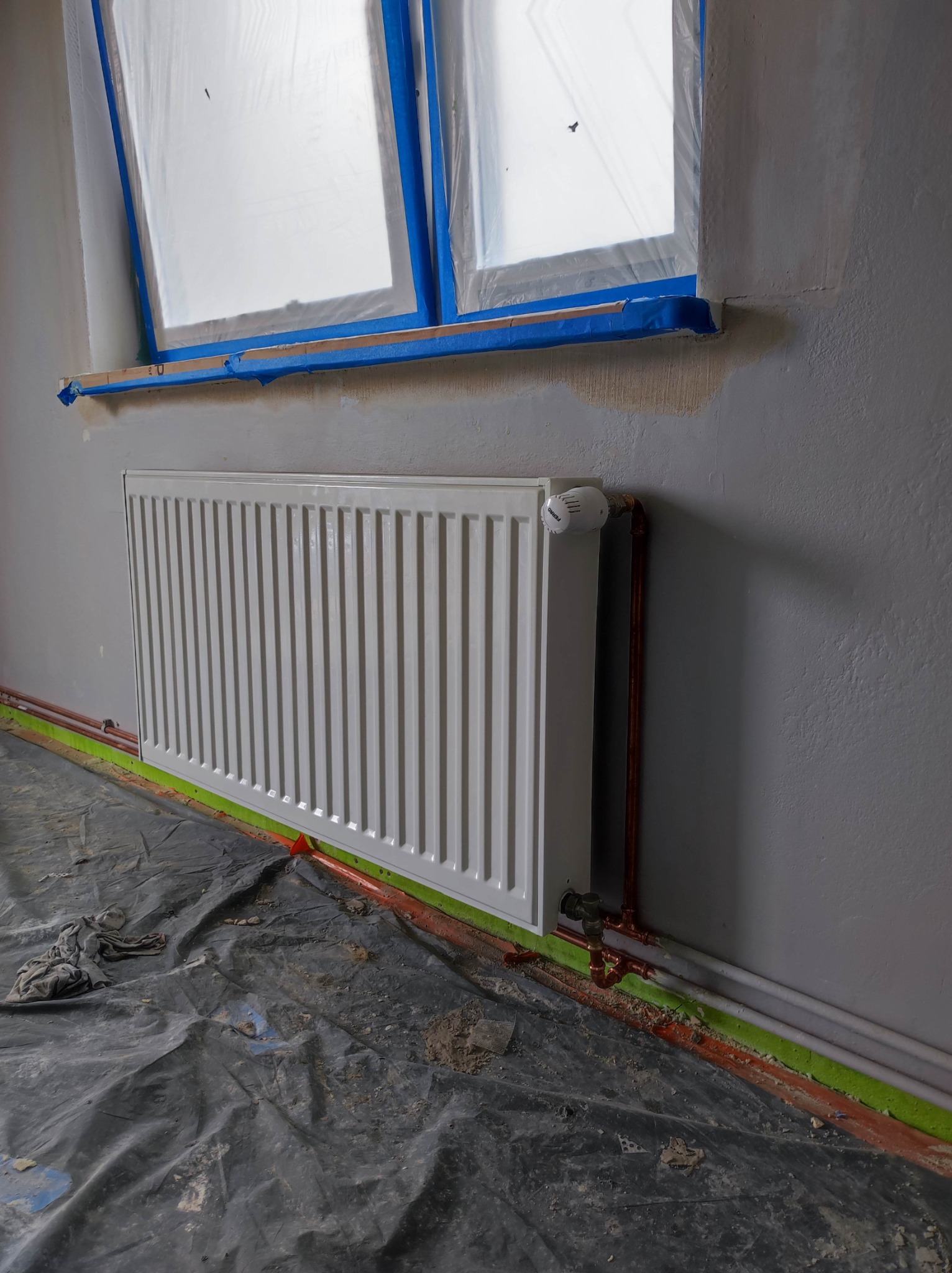 Nowy, biały grzejnik panelowy zamontowany na szarej ścianie w trakcie remontu, z widocznymi miedzianymi rurami i termostatem Ferro, okno zabezpieczone folią malarską.