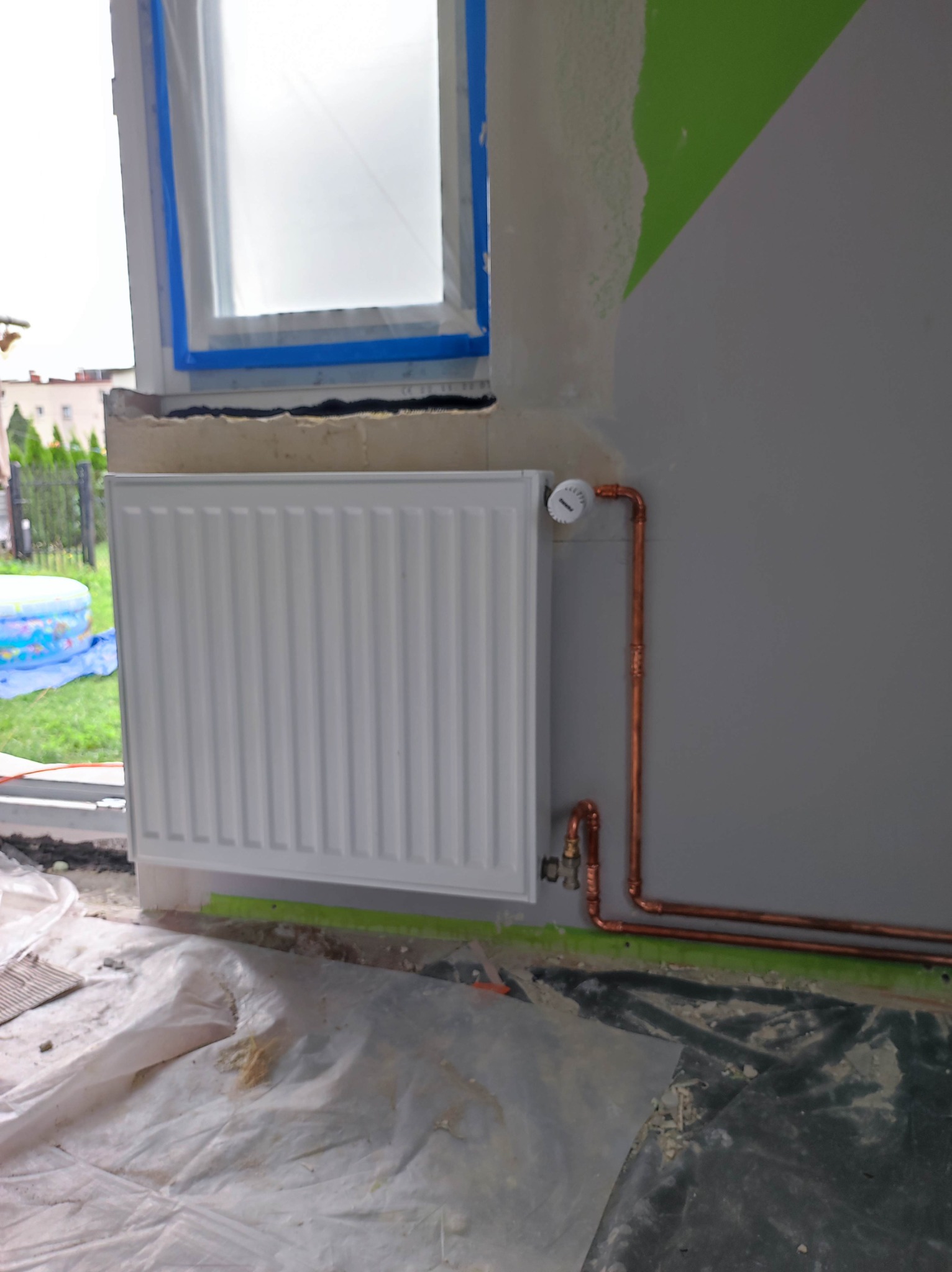 Biały grzejnik panelowy zamontowany pod oknem w trakcie prac wykończeniowych, widoczne miedziane rury instalacji grzewczej, folia ochronna na podłodze i fragment pomalowanej ściany.