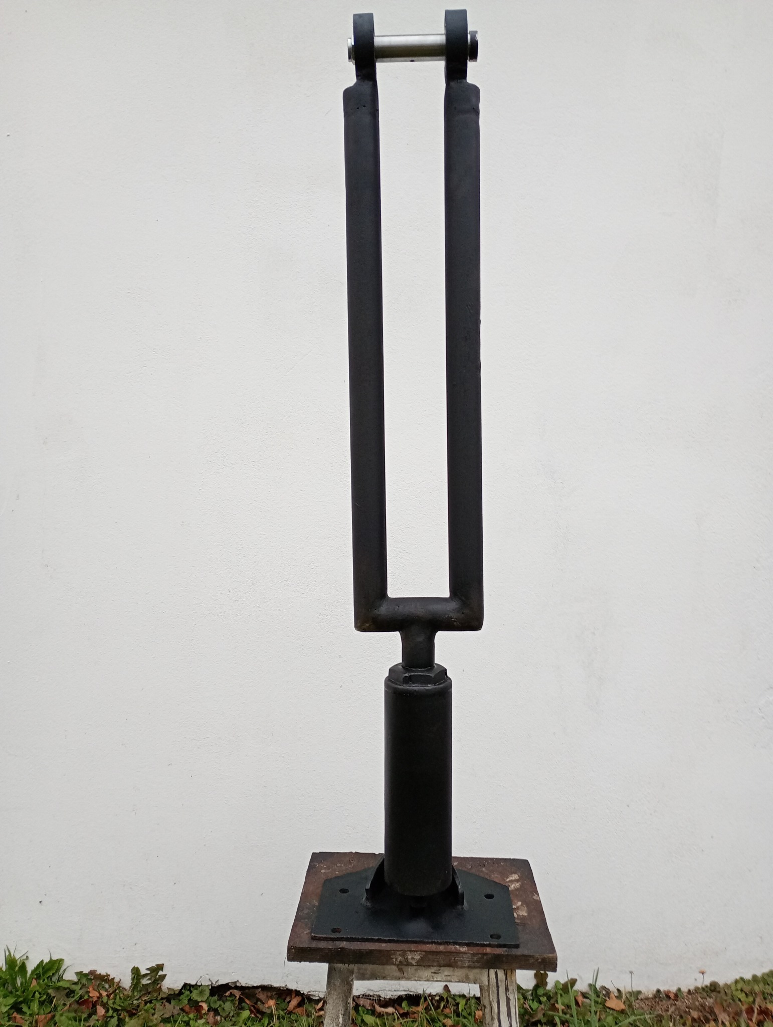 Spawana, czarna konstrukcja metalowa z dwoma pionowymi prętami połączonymi u góry poprzeczką, zamocowana na cylindrycznej podstawie przykręconej do metalowej płyty, całość ustawiona na niskim...