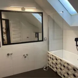 Nowoczesna łazienka na poddaszu z dużym lustrem w czarnej ramie, kabiną prysznicową ze szklanymi drzwiami i biało-czarną mozaiką na obudowie wanny.