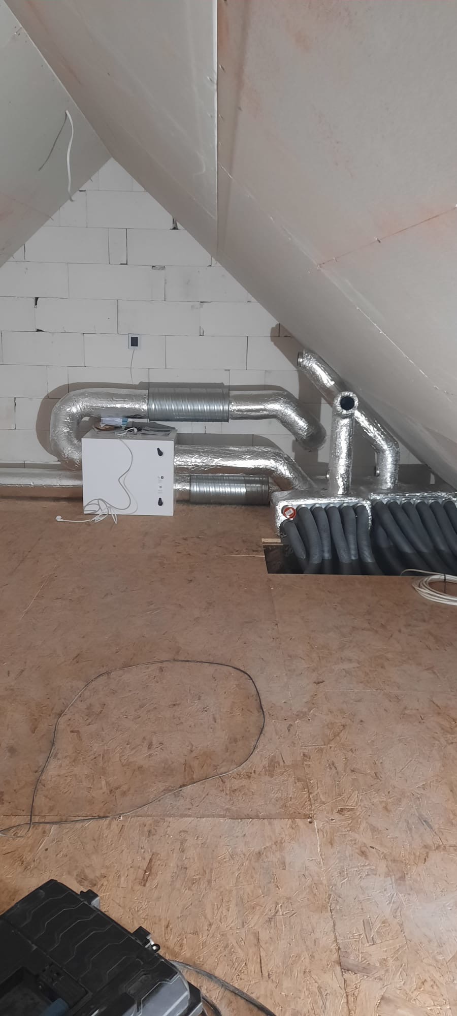 Instalacja rekuperacji na poddaszu: biała centrala wentylacyjna z podłączonymi srebrnymi rurami wentylacyjnymi, widoczne przewody wentylacyjne w izolacji termicznej, surowy stan wykończenia wnętrza.