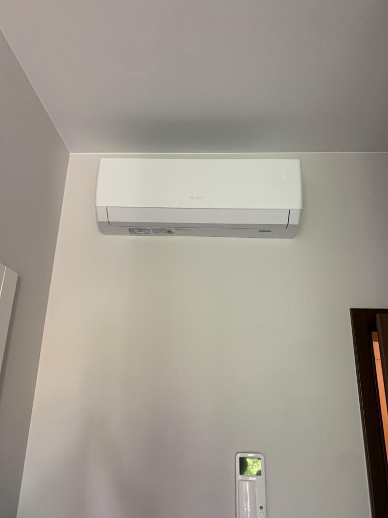 Biały klimatyzator ścienny z czerwoną diodą kontrolną, zamontowany w jasnym pomieszczeniu z szarą ścianą i fragmentem brązowej framugi drzwi w prawym dolnym rogu.