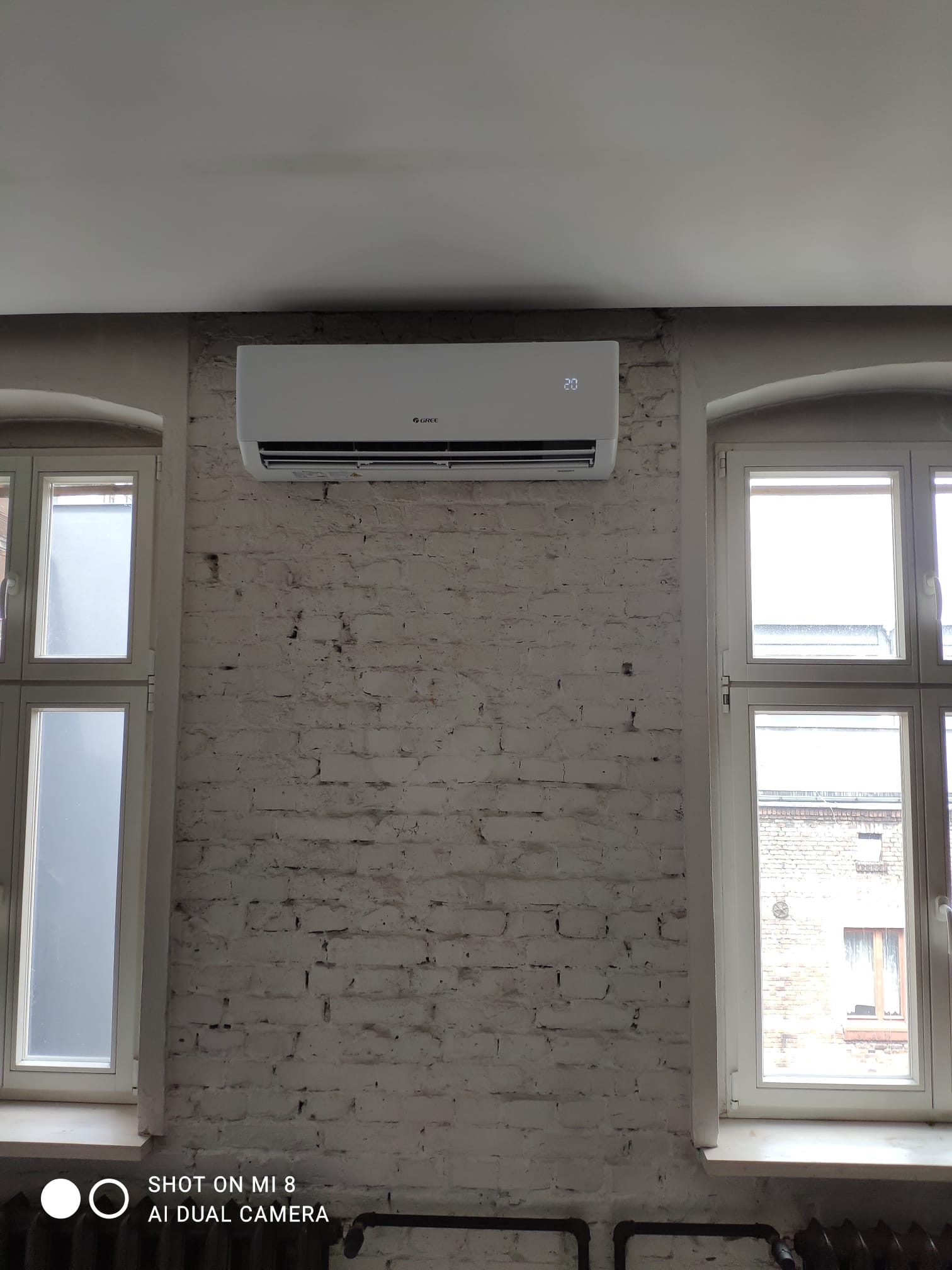 Klimatyzator marki Gree zamontowany na białej, ceglanej ścianie między dwoma oknami z widokiem na budynek.