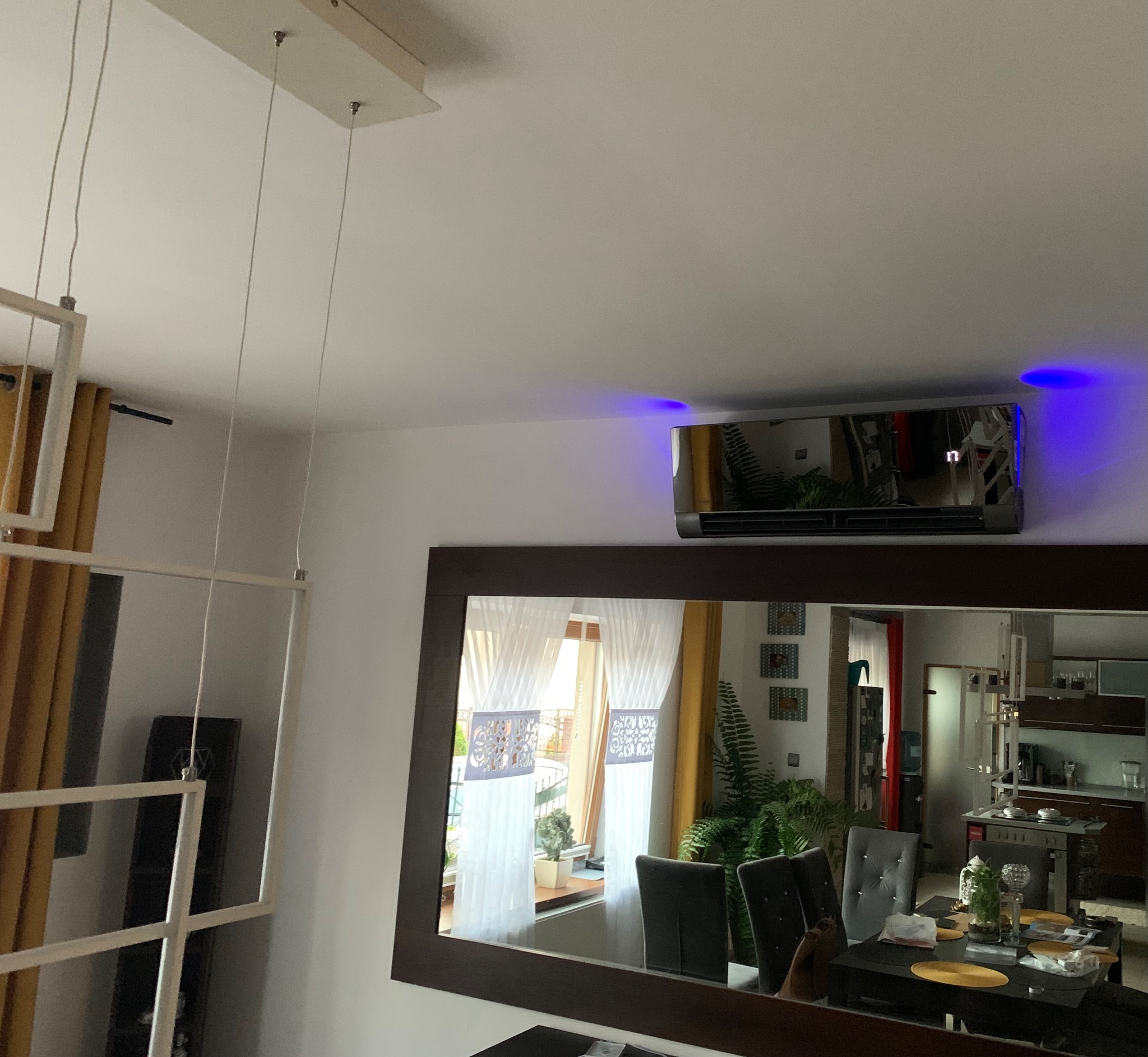 Nowoczesny klimatyzator ścienny z niebieskim podświetleniem LED, zamontowany nad dużym lustrem w jadalni, odbijającym widok okna, stołu i kuchni.