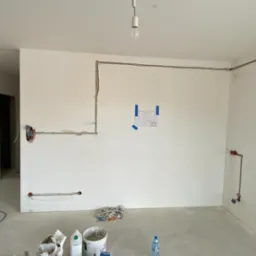 Surowy stan wnętrza w trakcie prac elektrycznych: widoczne przewody poprowadzone po ścianach, puszki elektryczne, plan pomieszczenia przyklejony taśmą, surowe ściany i podłoga, tymczasowe...