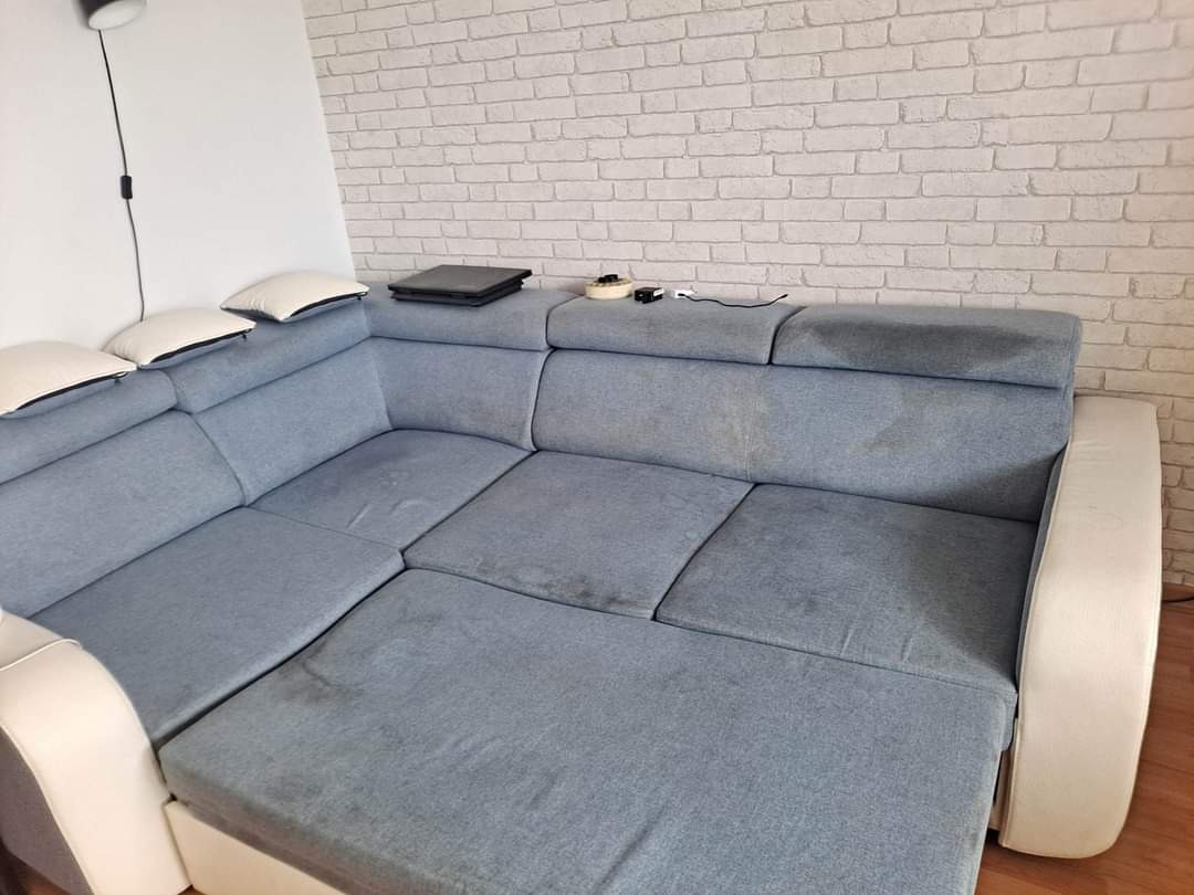 Szara, narożna kanapa z widocznymi plamami na siedzisku i oparciu, stojąca na tle ściany z cegły.