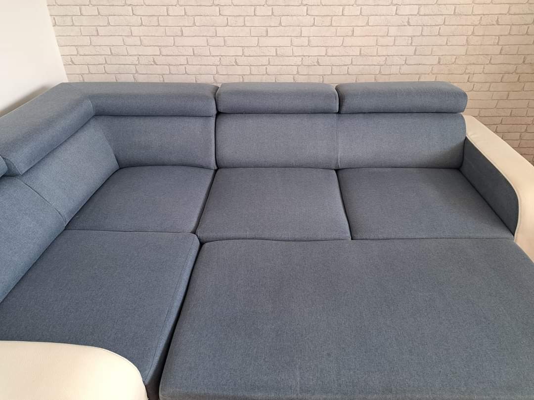 Narożnik z szarej tkaniny z regulowanymi zagłówkami i białymi elementami, na tle ściany z białej cegły.