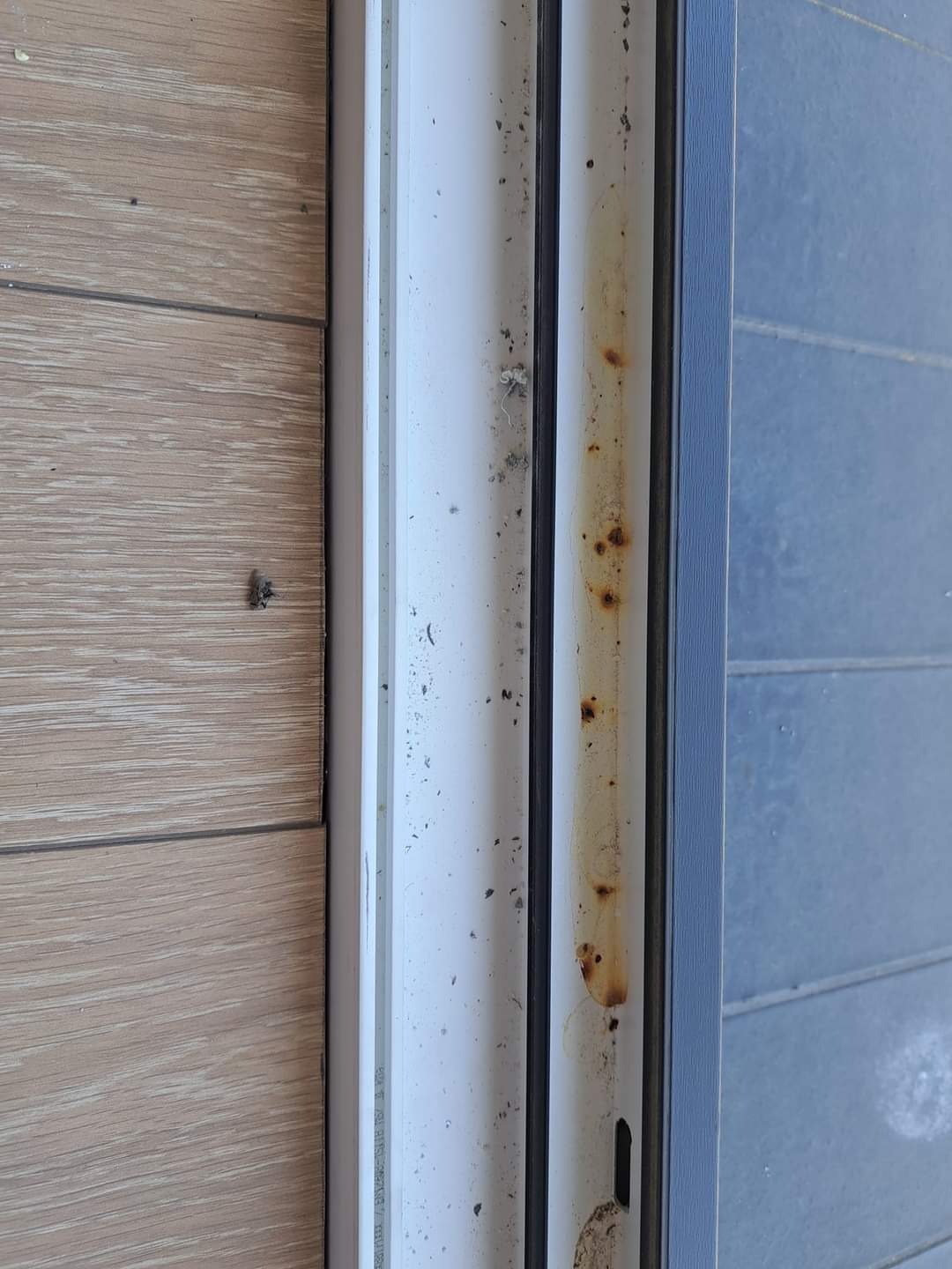 Silnie zabrudzona, biała rama okienna z widocznymi plamami rdzy i kurzu, sąsiadująca z drewnopodobną okładziną ścienną i niebieską ramą okna.