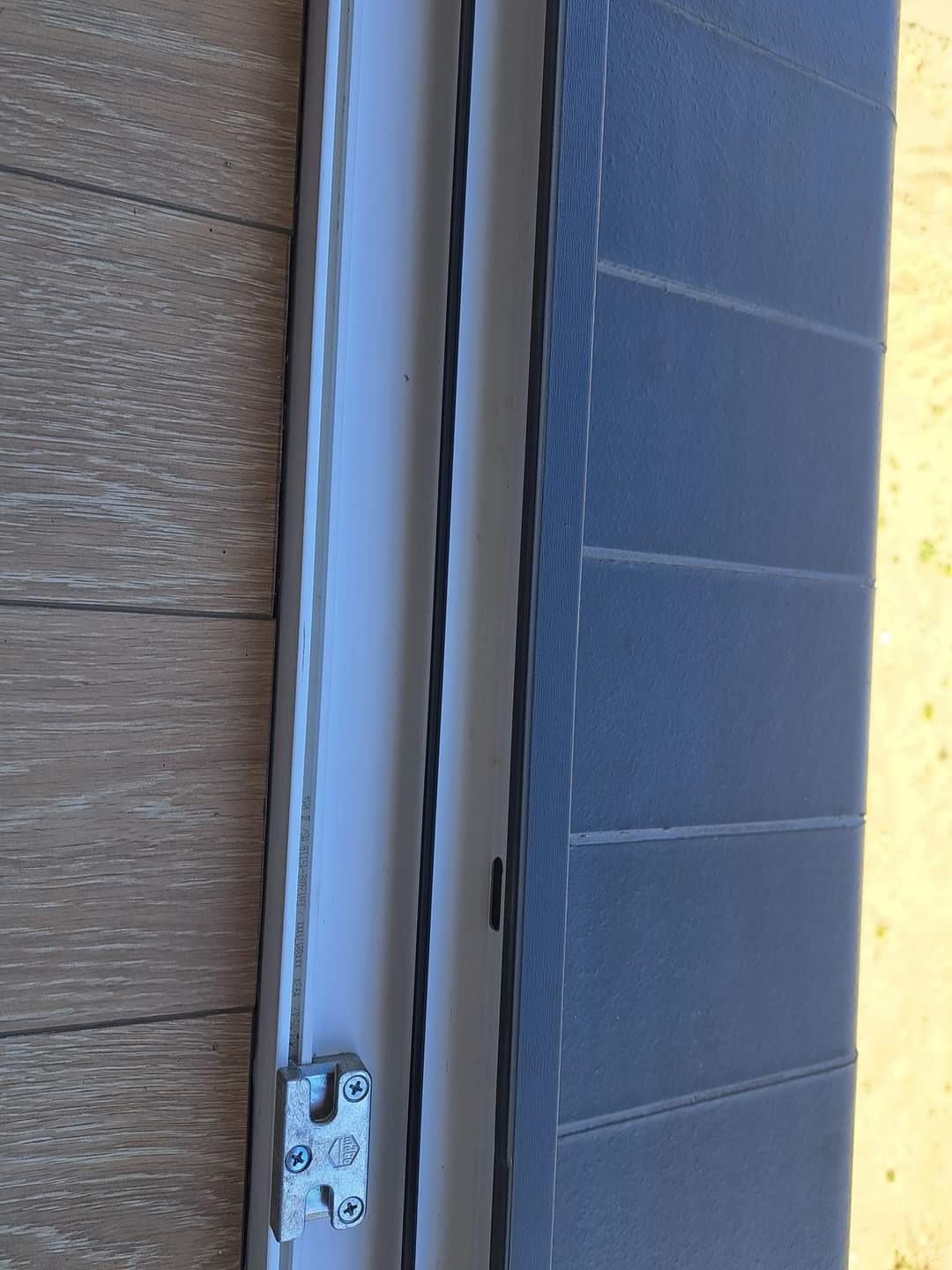 Fragment okna PCV z widocznym zamkiem, uszczelkami i elementami ramy, na tle drewnopodobnej okładziny i szarej ściany zewnętrznej.