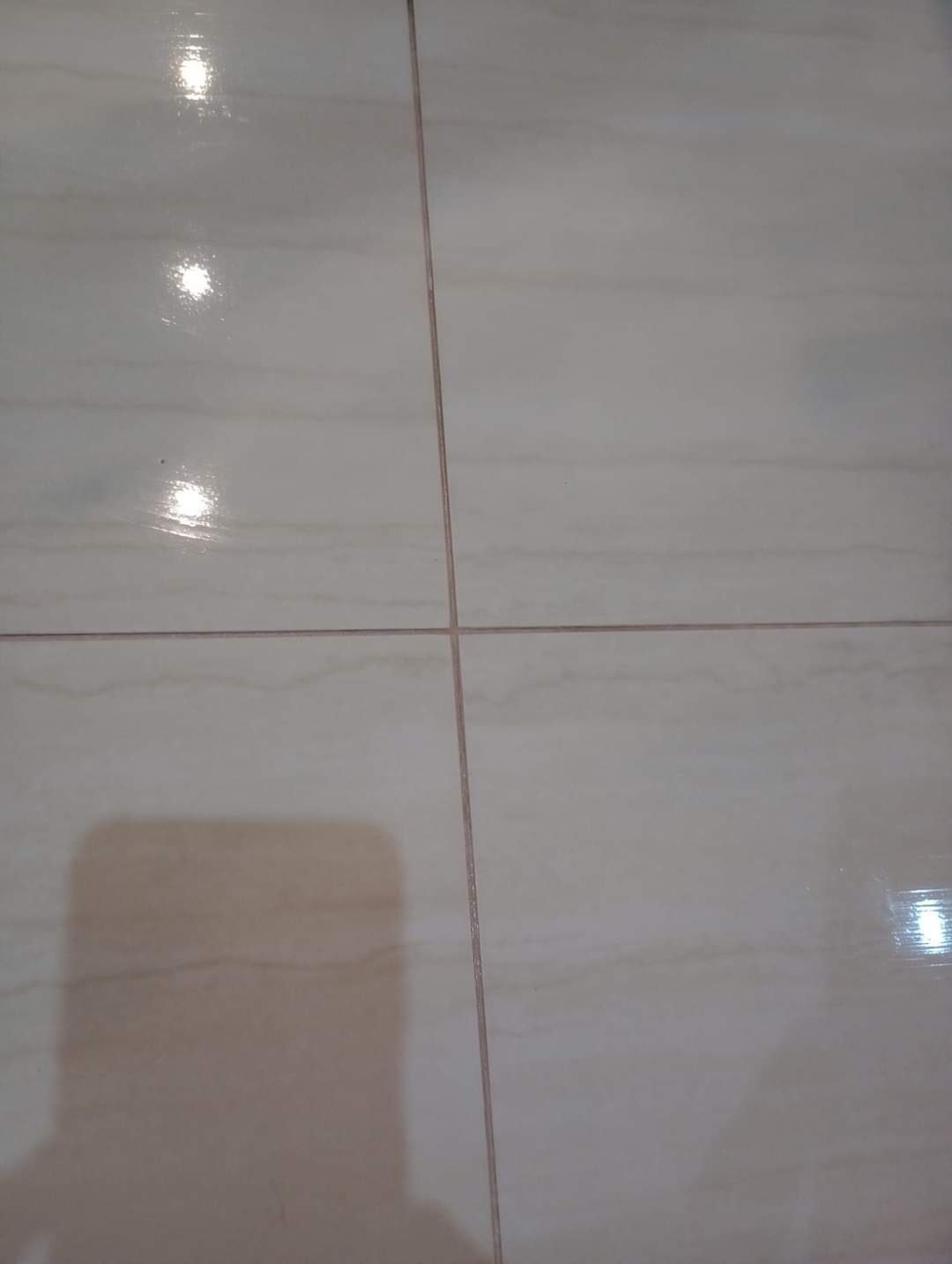 Błyszczące, beżowe płytki podłogowe z delikatnym wzorem marmuru i ciemniejszymi fugami, widoczne odbicia światła i cień osoby robiącej zdjęcie.