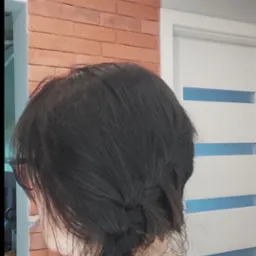 Kobieta z ciemnymi włosami zaplecionymi w warkocz, widoczny fragment ściany z cegły i białe drzwi z niebieskimi akcentami.