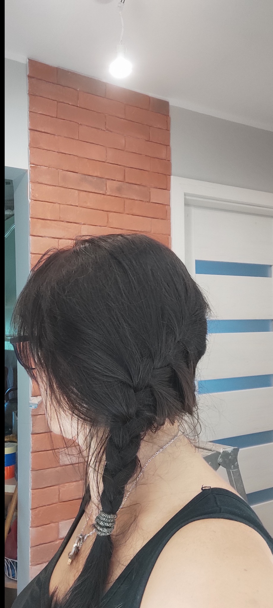 Kobieta z ciemnymi włosami zaplecionymi w warkocz, widoczny fragment ściany z cegły i białe drzwi z niebieskimi akcentami.