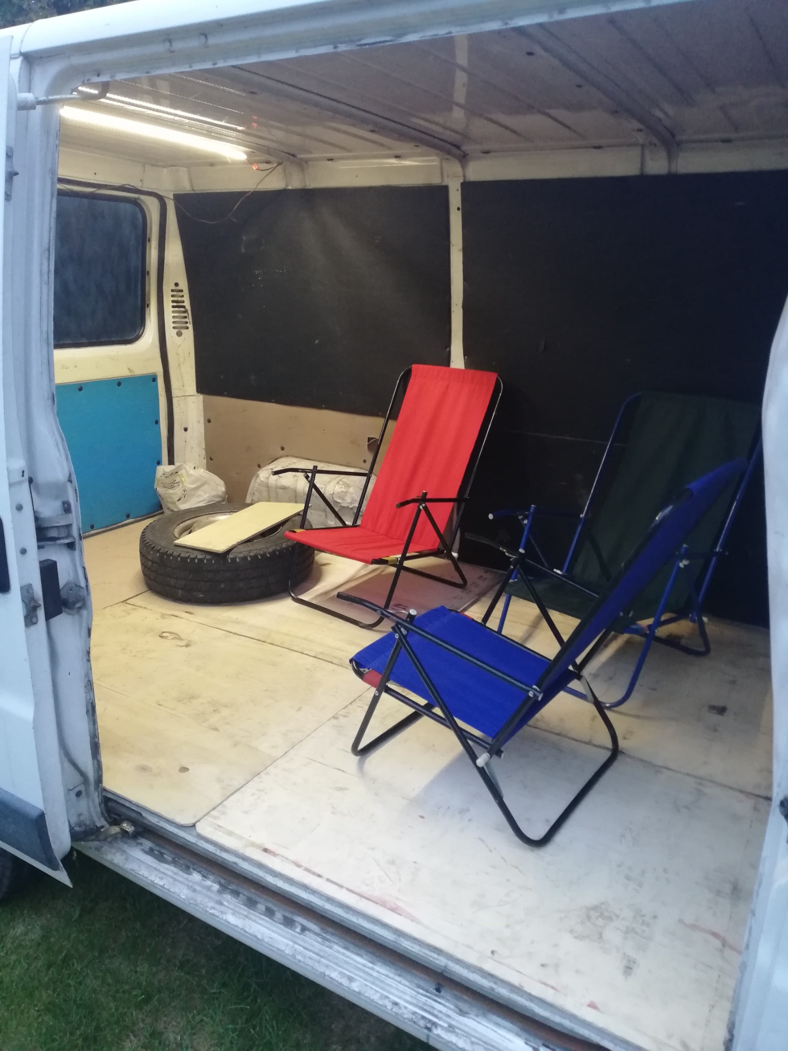 Wnętrze białego vana z otwartymi drzwiami bocznymi, w środku opona z deską, czerwone, niebieskie i zielone składane krzesła, podświetlane oświetleniem LED.