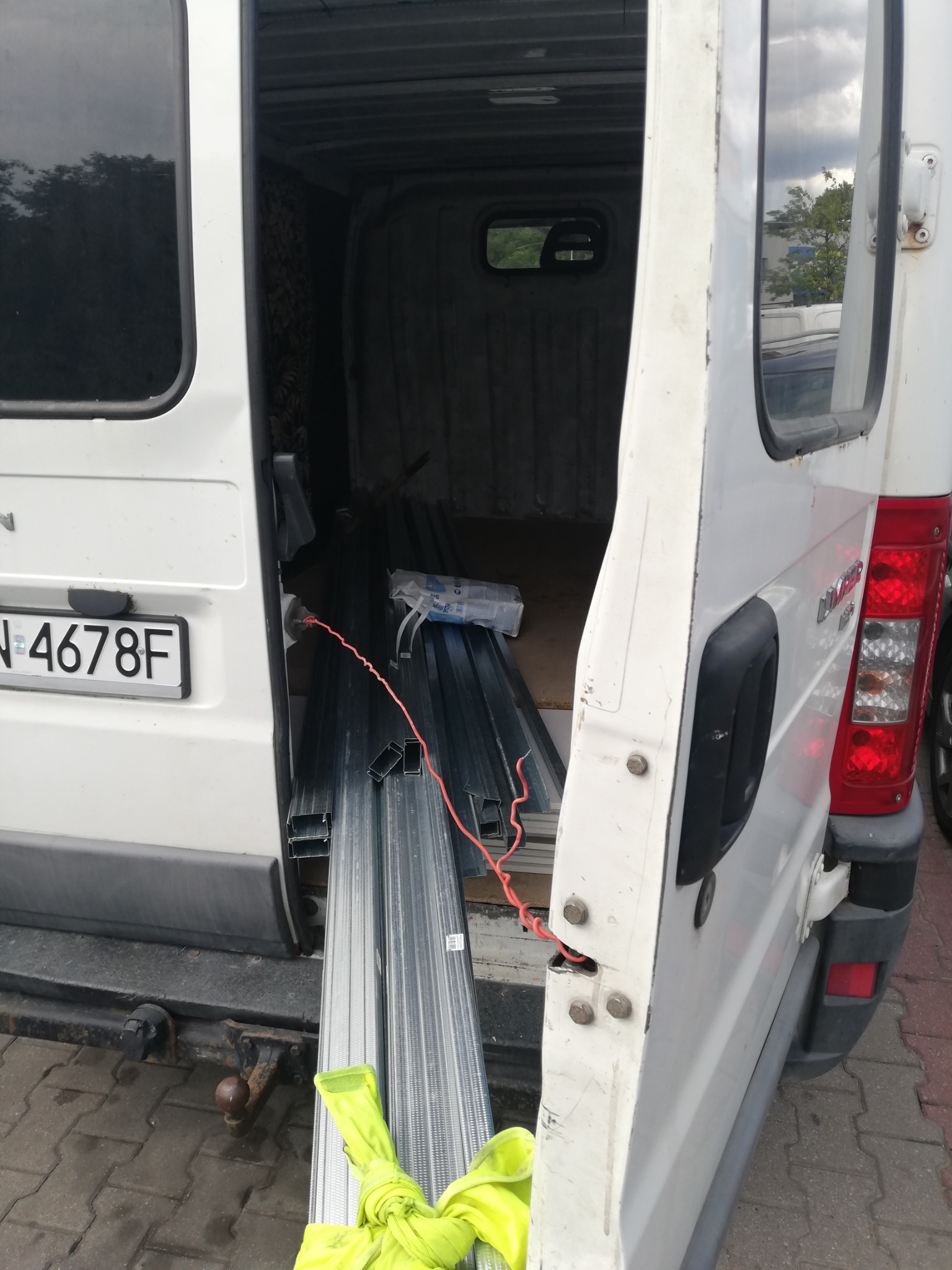 Wnętrze białego busa z otwartymi drzwiami, załadowane metalowymi profilami i żółtą tkaniną, widoczny hak holowniczy i tablica rejestracyjna.