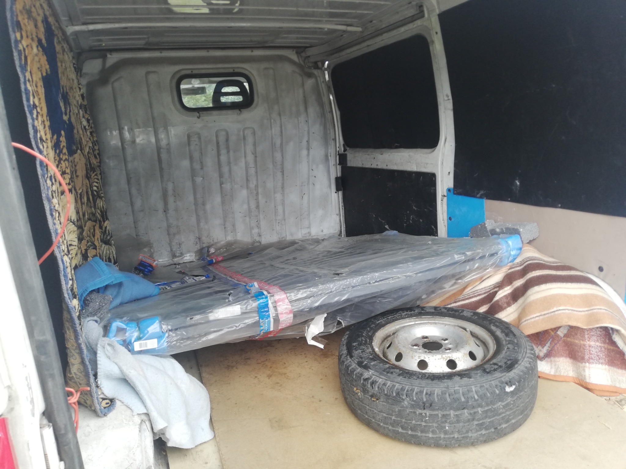 Wnętrze przestrzeni ładunkowej dostawczego samochodu: zapakowane w folię profile, opona, pled w paski i dywan z motywem roślinnym.