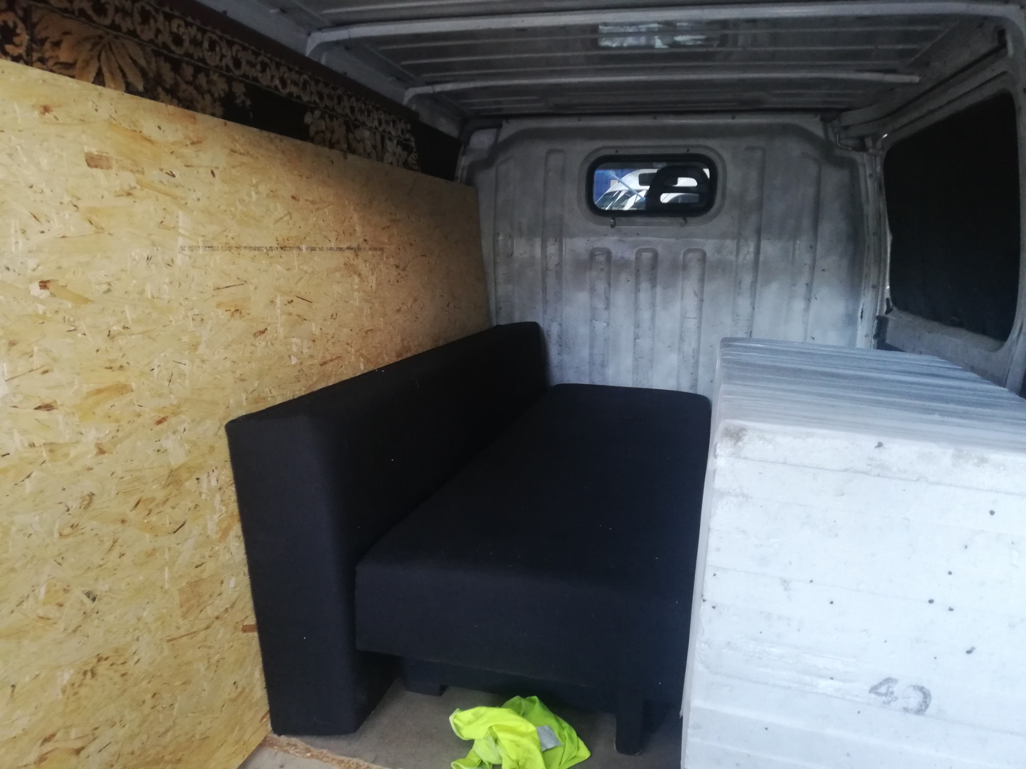 Wnętrze furgonetki z zabezpieczonymi meblami (czarna sofa) i płytami OSB, przygotowane do transportu. Widoczna odblaskowa kamizelka na podłodze.