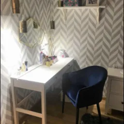 Biała toaletka z szufladami, na blacie kosmetyki i akcesoria do makijażu, nad nią geometryczne lustro i wiszące lampy, w tle tapeta w szare zygzaki, obok ciemnoniebieski, welurowy fotel.