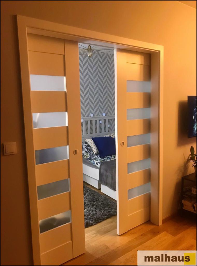 Rozsuwane drzwi wewnętrzne z przeszkleniami w kolorze ciepłego beżu, widok na sypialnię z łóżkiem i wzorzystą tapetą w tle.