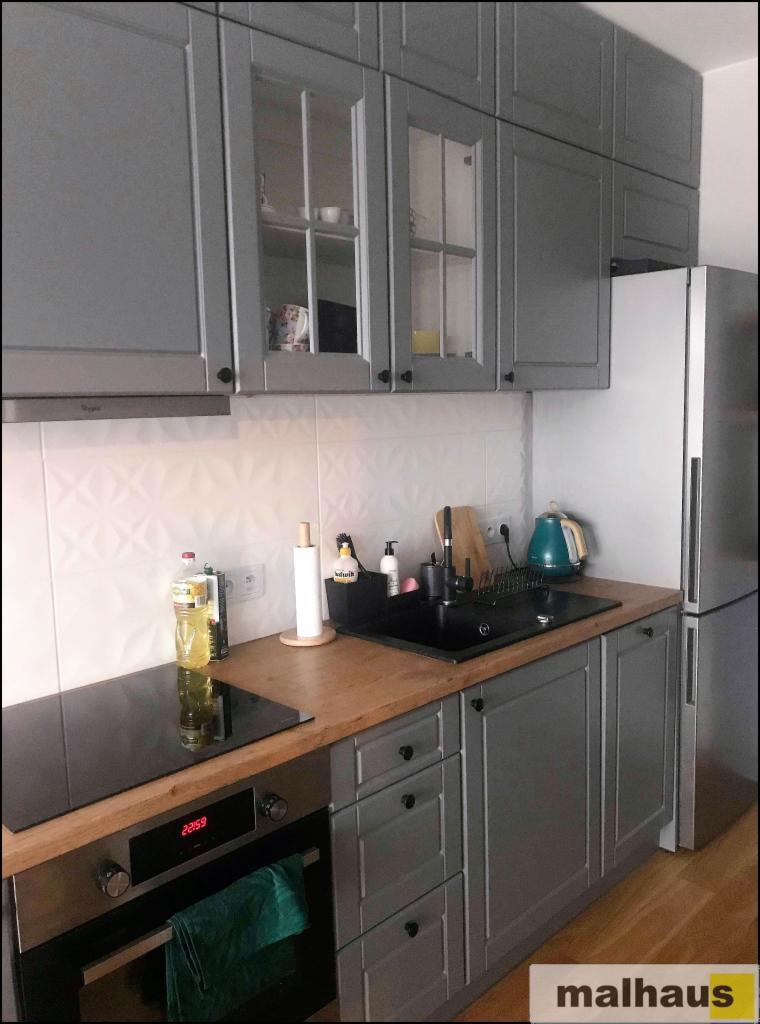 Szara zabudowa kuchenna z drewnianym blatem, czarnym zlewem i płytą indukcyjną. Na blacie widoczne akcesoria kuchenne i butelka oleju, a w tle białe, wzorzyste płytki.