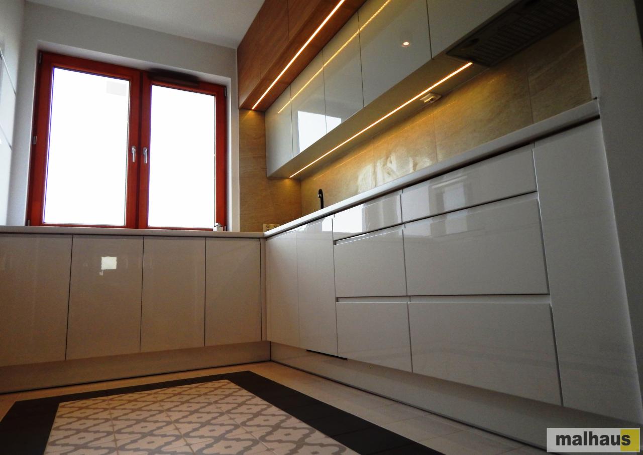 Nowoczesna kuchnia z białymi, lakierowanymi frontami szafek, drewnianym panelem nad górnymi szafkami z oświetleniem LED i wzorzystą podłogą.