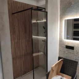 Nowoczesna kabina prysznicowa z czarnymi profilami, drewniane panele ścienne, podświetlane lustro i dekoracyjne płytki w wykończonej łazience. Na krześle leży karton z elementami montażowymi.