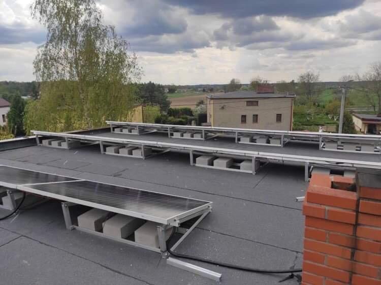 Montaż paneli słonecznych na płaskim dachu z widocznymi betonowymi blokami pod konstrukcją wsporczą i kominem z czerwonej cegły w prawym dolnym rogu, w tle budynki i drzewa.