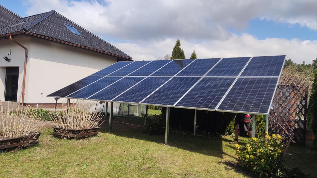 Instalacja paneli słonecznych na stelażu w ogrodzie przydomowym, widoczne panele, konstrukcja wsporcza, część domu z brązową dachówką oraz fragment osoby w tle.