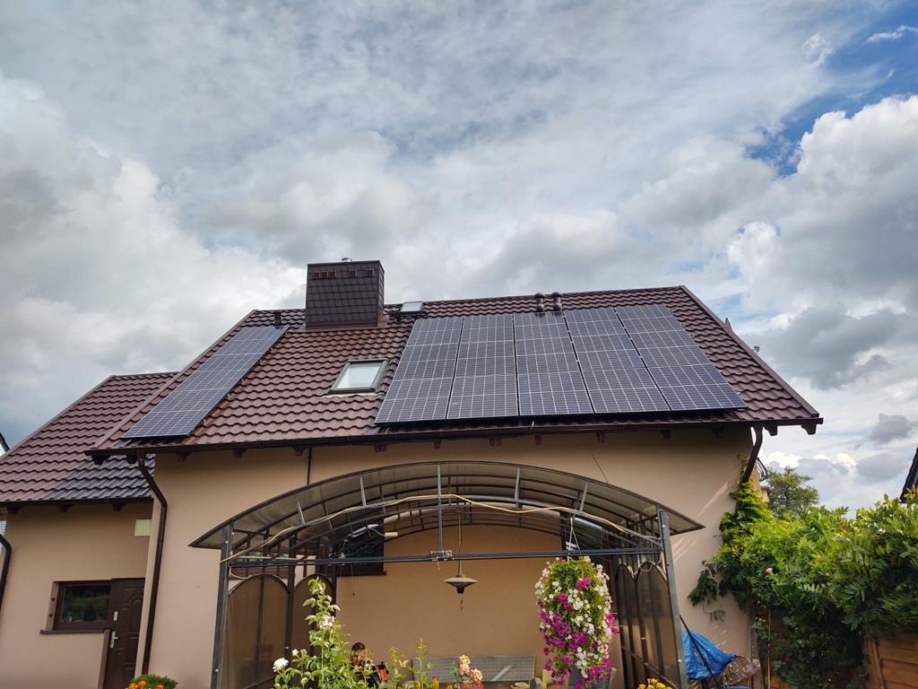 Dom jednorodzinny z brązowym dachem i panelami słonecznymi, widok z dołu na tle pochmurnego nieba.