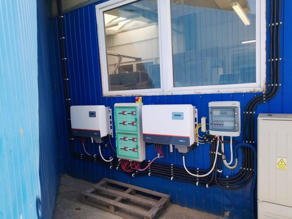 Instalacja elektryczna z dwoma falownikami i rozdzielnicami na niebieskiej ścianie z blachy trapezowej, widoczne kable w peszlach i paleta pod spodem.