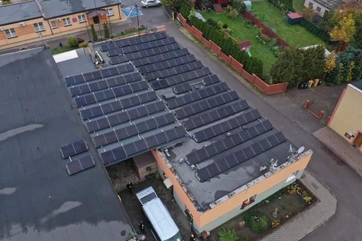 Widok z góry na dach budynku z zainstalowanymi panelami fotowoltaicznymi; widoczny zaparkowany bus serwisowy i pracownicy.