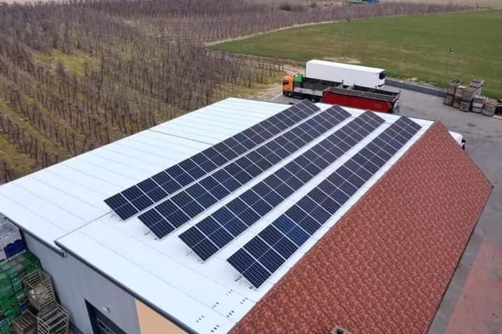 Panele słoneczne zamontowane na dachu budynku gospodarczego z czerwonymi dachówkami, na tle sadu i zaparkowanych ciężarówek.